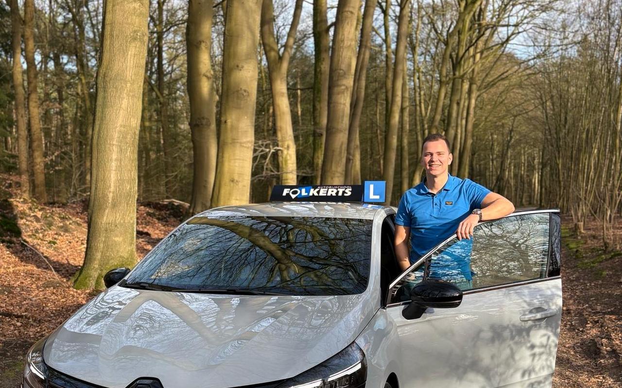Niek Folkerts naast de lesauto van Autorijschool Folkerts, een grijze Renault Clio met lesbord op een bospad